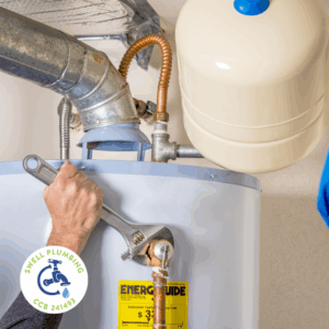 Plumber tightening fittings on a residential water heater during fall maintenance in Salem, OR, ensuring safe winter operation. Swell Plumbing logo shown in the lower corner.